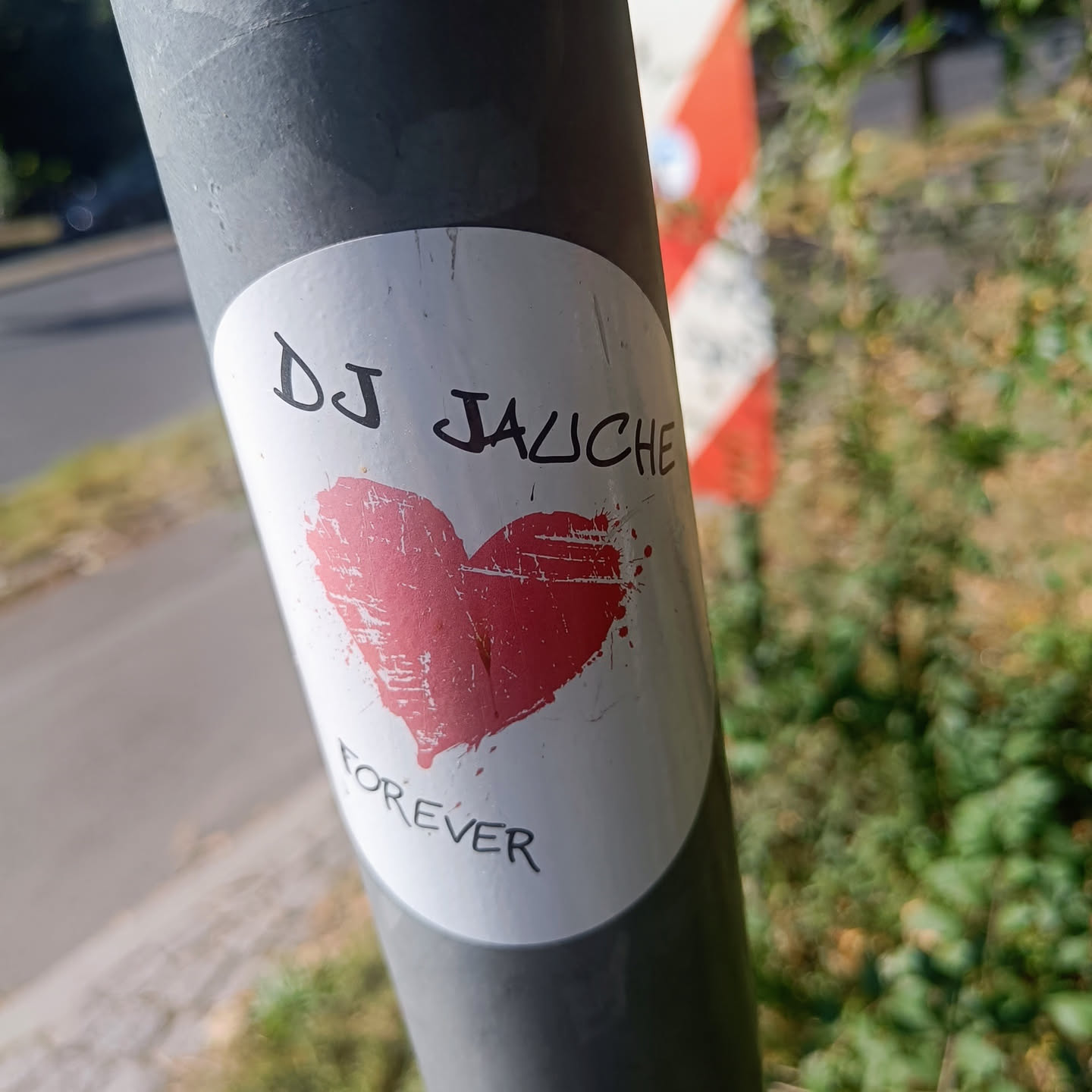 A round white sticker with a pink heart and the text 'DJ JAUCHE' on top and 'FOREVER' at the bottom. The heart is hand-drawn with a distressed texture. The sticker is affixed to a gray metal pole.