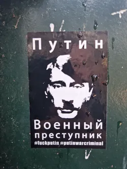 Black and white sticker depicting Vladimir Putin's face with text in Russian and English.  The Russian text translates to "Putin - War Criminal". The English hashtags further emphasize this.