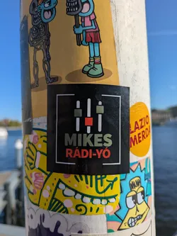 A square sticker featuring the logo for Mikes Rádió. The logo includes an image of equalizer bars and the text "MIKES RáDI-YÓ". The sticker is adhered to a light colored pole alongside other stickers.