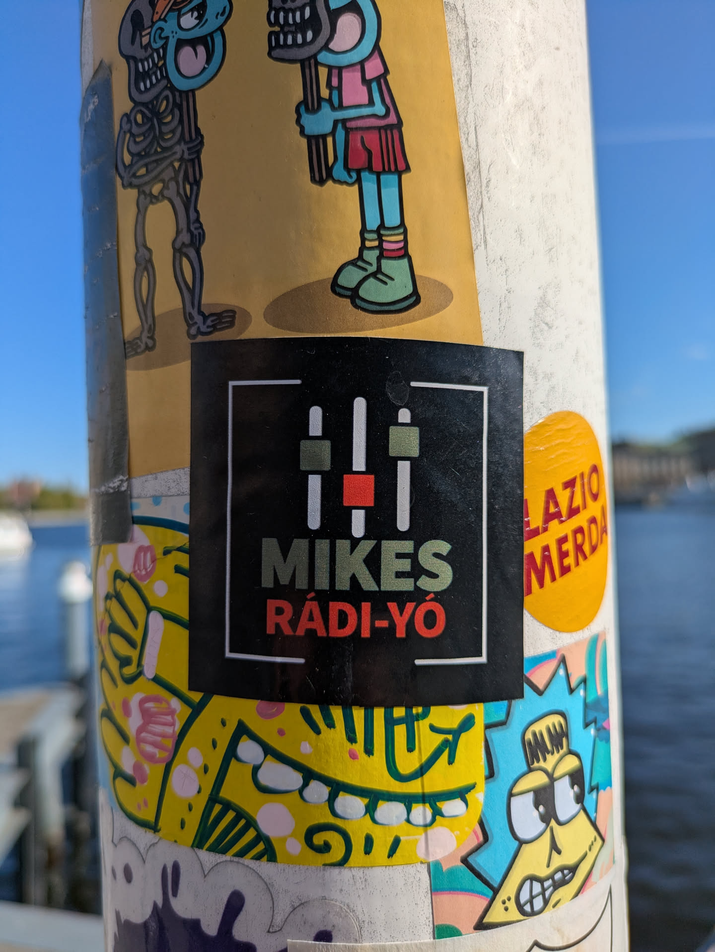 A square sticker featuring the logo for Mikes Rádió. The logo includes an image of equalizer bars and the text "MIKES RáDI-YÓ". The sticker is adhered to a light colored pole alongside other stickers.