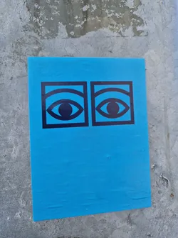 Bright blue sticker with a minimalist design featuring two eyes.  The eyes are black and outlined in a bold black square. The sticker is affixed to a concrete wall.
