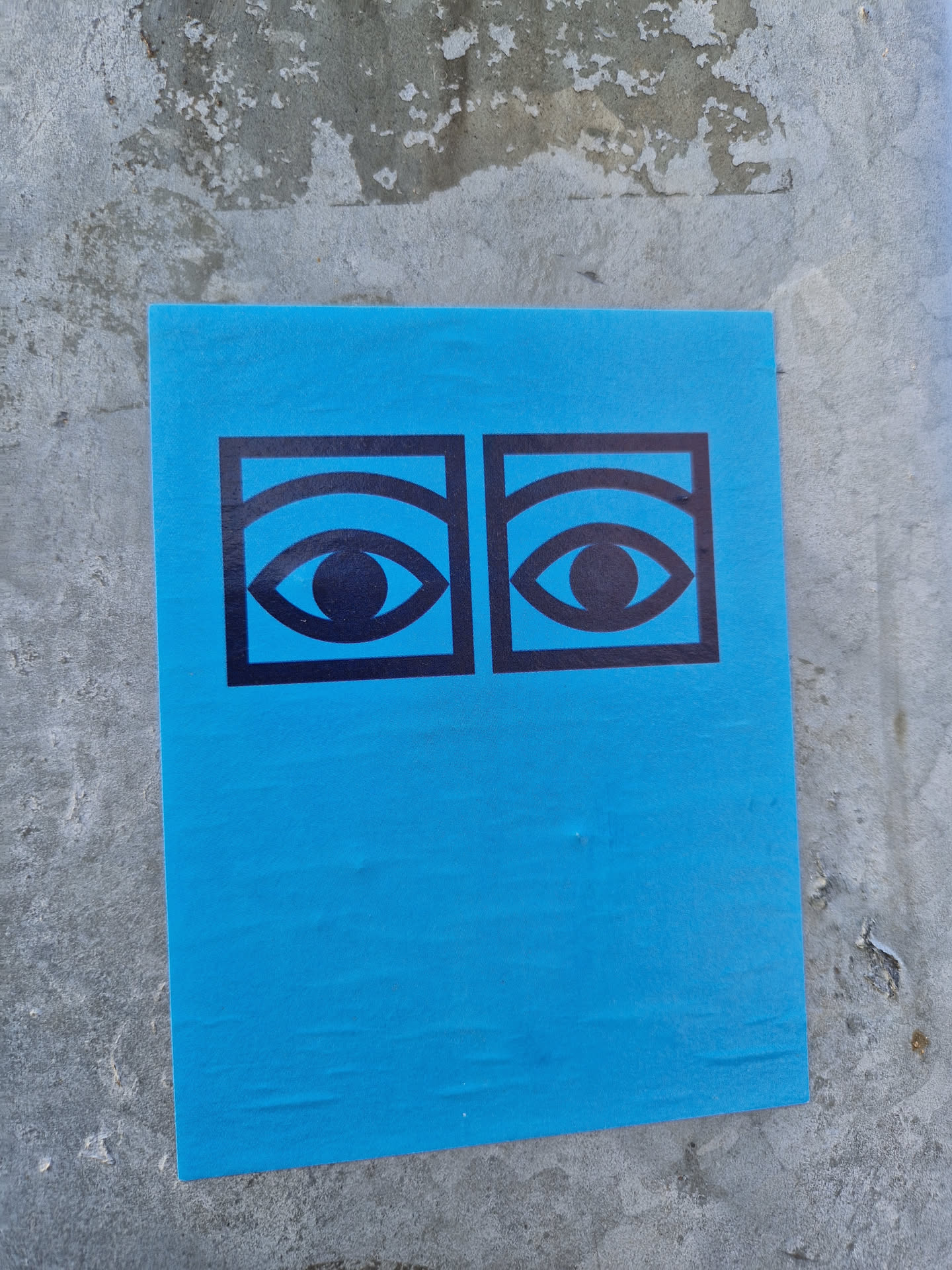 Bright blue sticker with a minimalist design featuring two eyes.  The eyes are black and outlined in a bold black square. The sticker is affixed to a concrete wall.
