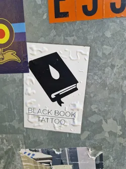 A sticker featuring a minimalist design of a book with a single teardrop, and the text "Black Book Tattoo" underneath.