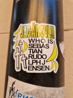 A sticker with a yellow and black design. It has the text "JAGÄR GUL!" at the top, followed by "Who is Sebastian Rudo LPHJ Ensen".  There are also images of bananas and crosses on the sticker.