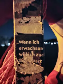 A worn sticker with a German phrase partially visible. The full sentence is unclear due to damage. The sticker is affixed to a pole outdoors at night.