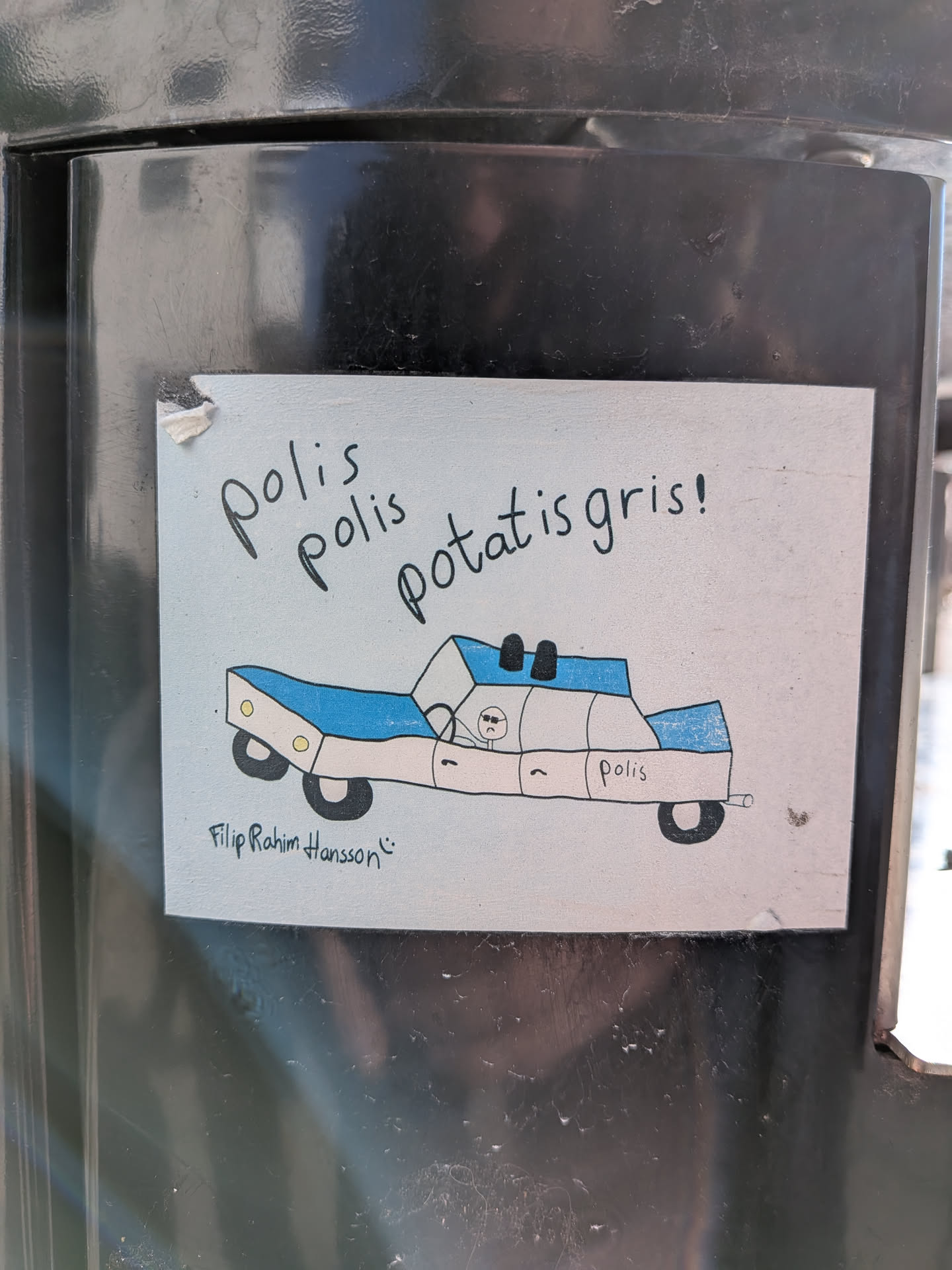 The sticker is rectangular with a light blue background. At the top, in black handwritten text, it reads 'polis polis potatis gris!'. Below the text is a hand-drawn image of a police car. The car is predominantly white with blue accents on the roof and sides. It has black wheels and yellow circular details on the front. A small, stick-figure-like character with sunglasses is drawn inside the car. The word 'polis' is written on the side of the car. Below the car, in smaller cursive handwriting, is 'Filip Rahim Hansson' followed by a smiley face emoji. The sticker appears to be slightly worn at the edges.