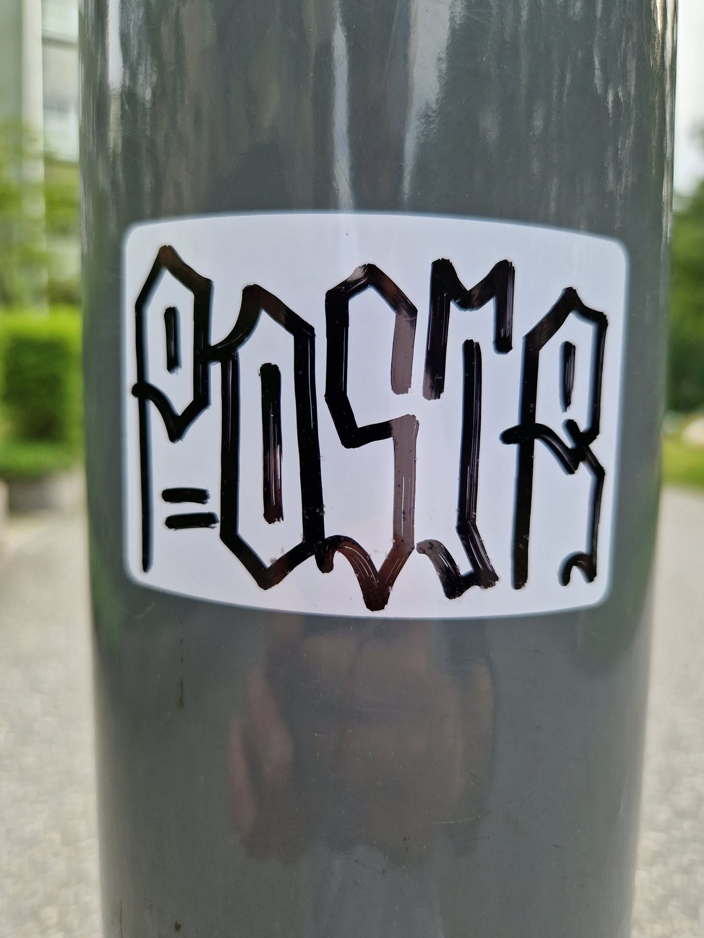 A white rectangular sticker with black stylized lettering that says "POSCA" in a bold, graffiti-inspired font. The sticker is affixed to a gray pole outdoors.