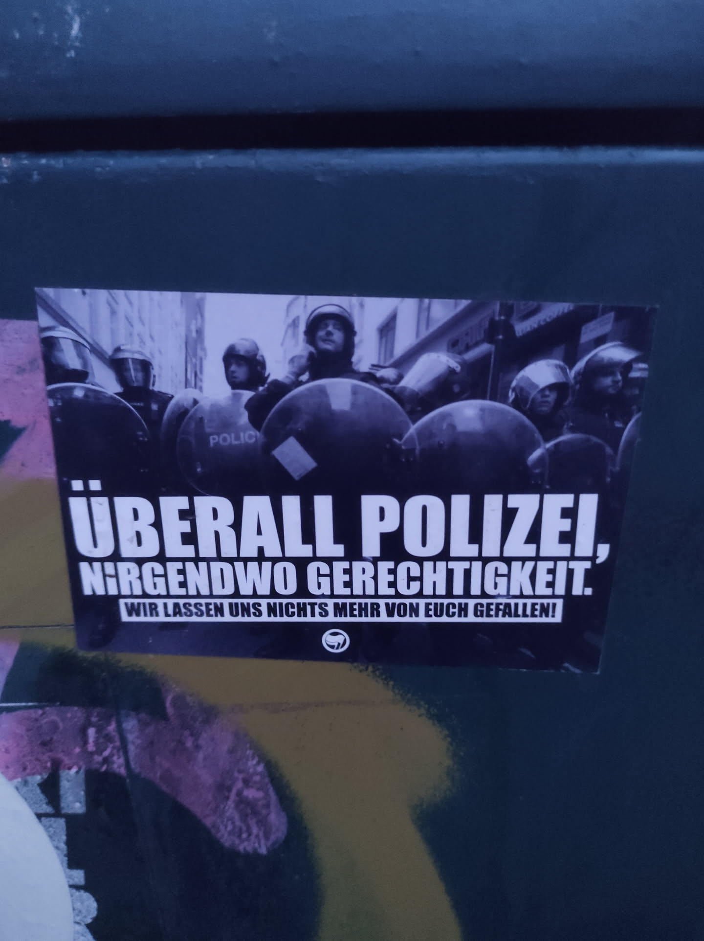 A sticker depicting a line of riot police with the text "ÜBERALL POLIZEI, NIRGENDWO GERECHTIGKEIT. WIR LASSEN UNS NICHTS MEHR VON EUCH GEFALLEN!" which translates to: "Police everywhere, justice nowhere. We won't let you get away with anything anymore!"
