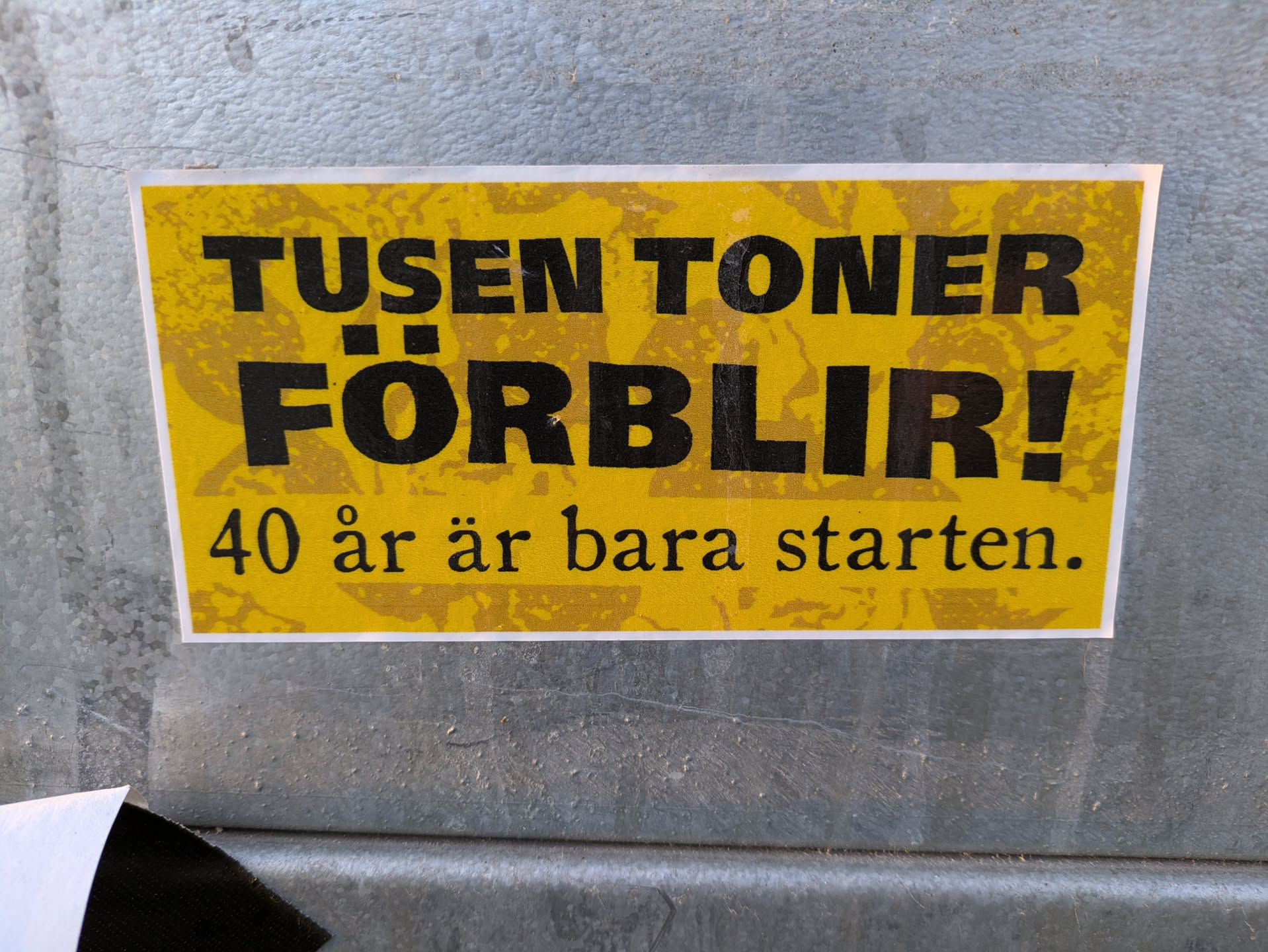 Yellow sticker with black text.  The text says "Tusen Töner Förlir!" which translates from Swedish to "A Thousand Tones Remain!"  Below it, in smaller text, it says "40 år är bara starten." which translates to "40 years is just the beginning."