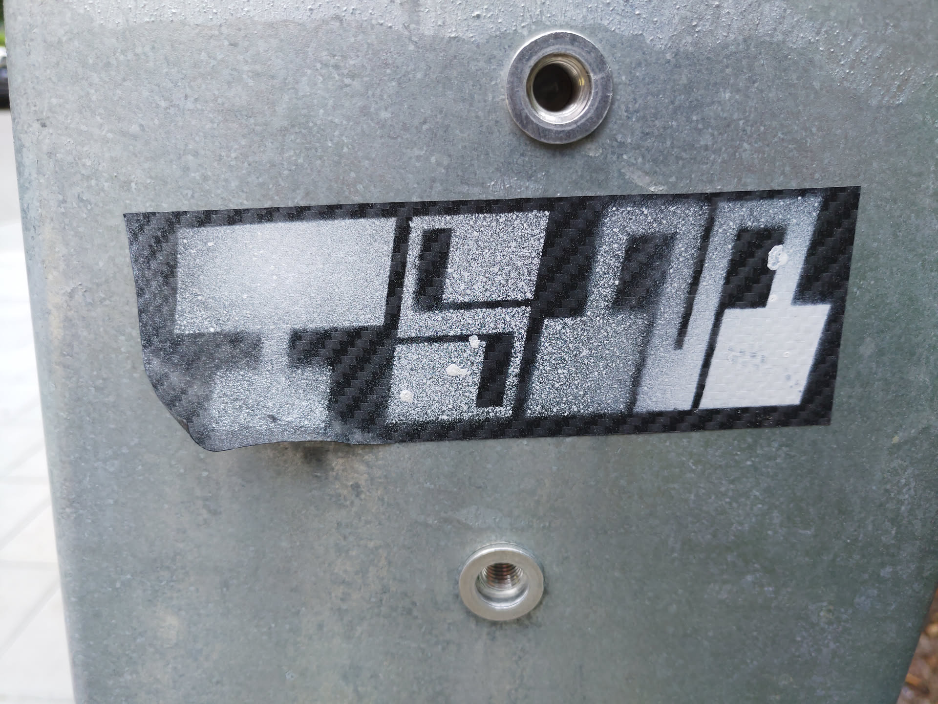 A black and white sticker with an abstract design, possibly representing buildings or structures. It's applied to a metallic surface. The sticker has a carbon fiber-like texture.