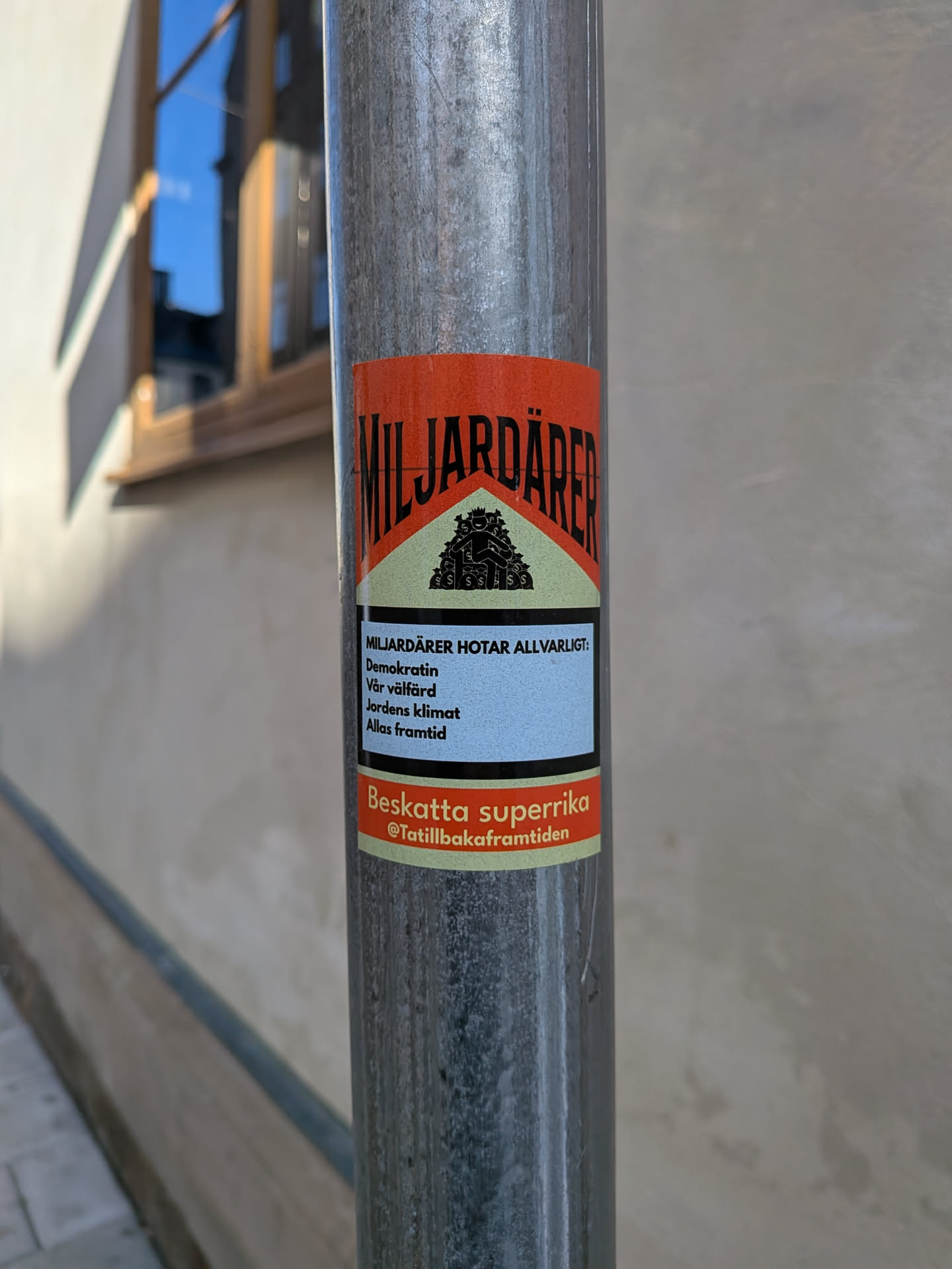 A rectangular sticker adhering to a metal pole, designed as a parody of a Marlboro cigarette pack. The top red chevron features the bold black text 'MILJARDÄRER' above a black silhouette emblem showing a crowned figure sitting atop bags of money. The middle section contains a warning label styled box with black borders reading: 'MILJARDÄRER HOTAR ALLVARLIGT: Demokratin, Vår välfärd, Jordens klimat, Allas framtid'. The bottom red and off-white section features the text 'Beskatta superrika' followed by the handle '@Tatillbakaframtiden'.