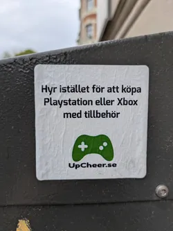 A square sticker is adhered to a metal surface. The sticker is white with black text and a green game controller icon. It reads 'Hyr istället för att köpa Playstation eller Xbox med tillbehör' and 'UpCheer.se'. The sticker promotes renting gaming consoles from UpCheer.se.