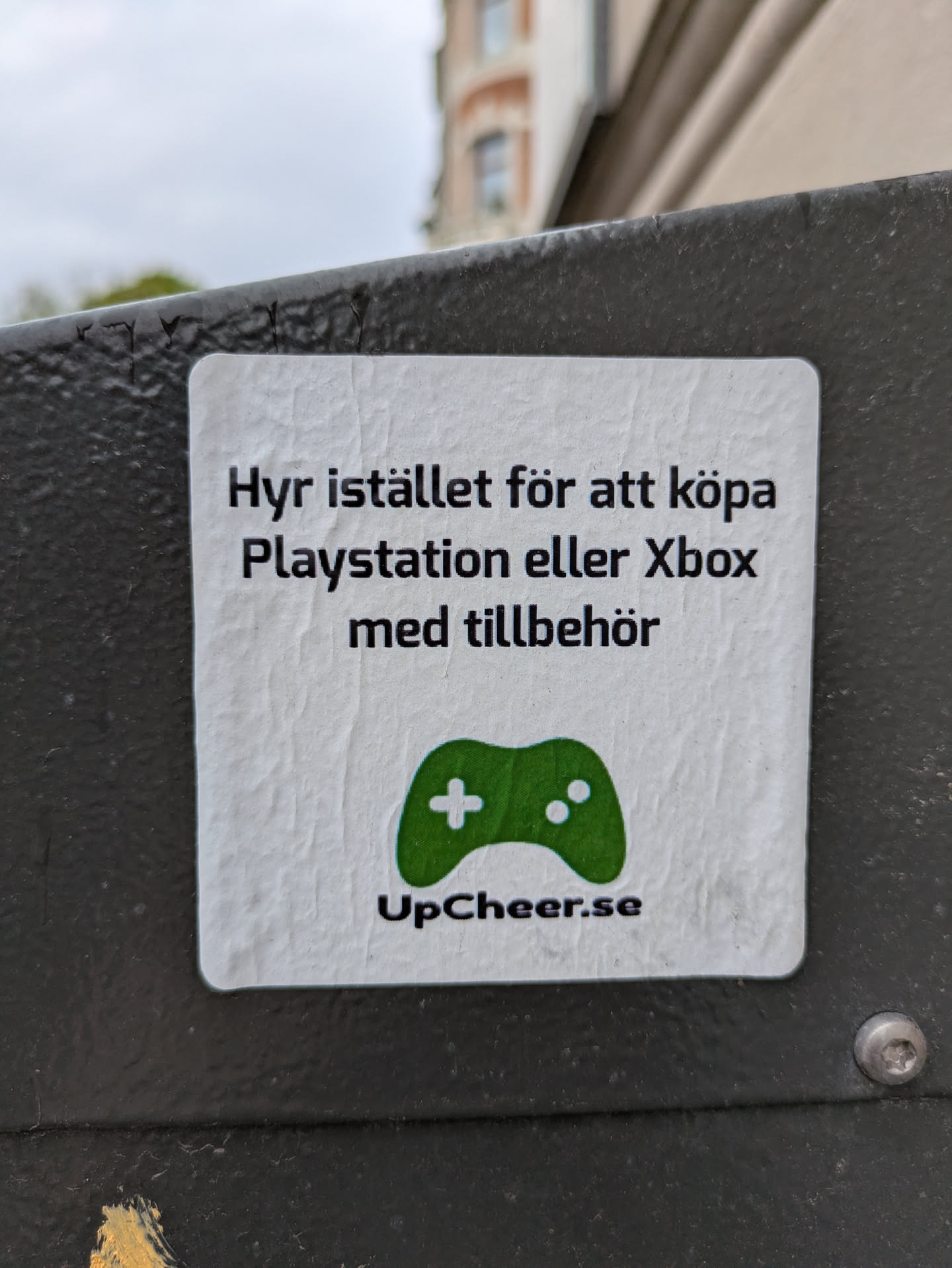 A square sticker is adhered to a metal surface. The sticker is white with black text and a green game controller icon. It reads 'Hyr istället för att köpa Playstation eller Xbox med tillbehör' and 'UpCheer.se'. The sticker promotes renting gaming consoles from UpCheer.se.