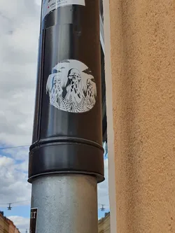 A round sticker featuring a minimalist design of three cloaked figures under a cloudy sky. The style is reminiscent of occult or surreal art, evoking a sense of mystery and enigma.  The sticker is affixed to a dark-colored pipe.
