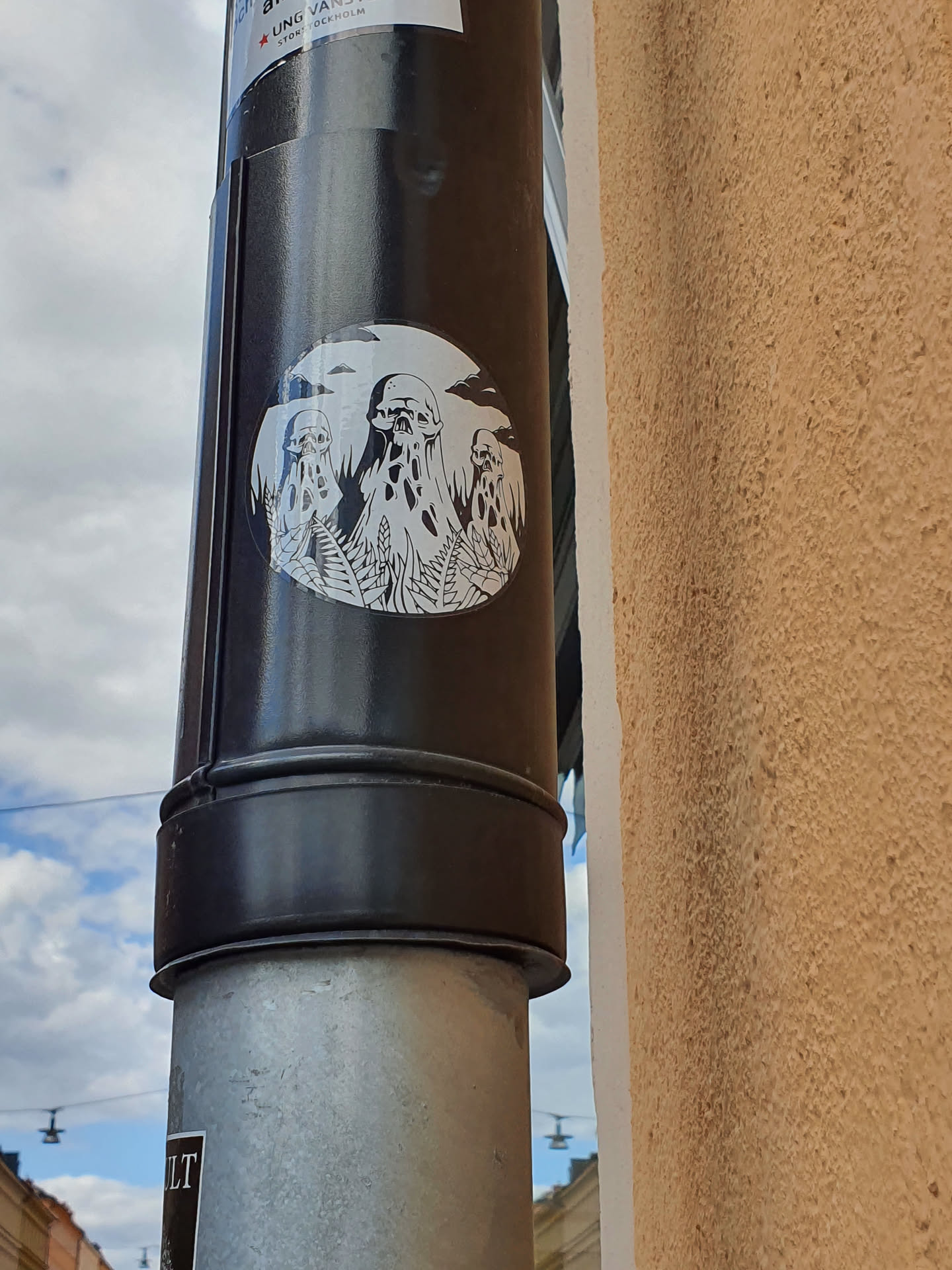 A round sticker featuring a minimalist design of three cloaked figures under a cloudy sky. The style is reminiscent of occult or surreal art, evoking a sense of mystery and enigma.  The sticker is affixed to a dark-colored pipe.