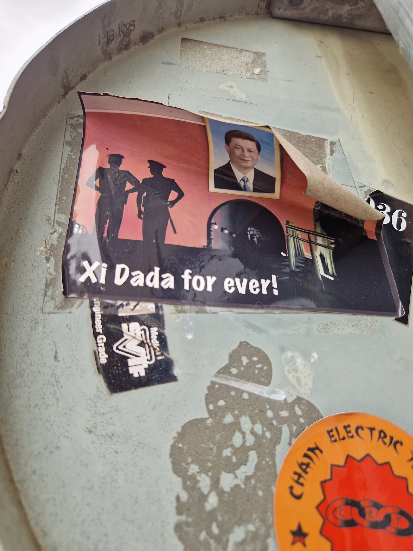A sticker featuring a stylized image of Xi Jinping, the current paramount leader of China. The sticker also includes two silhouetted figures in military-style uniforms and what appears to be a tunnel or gate. The overall aesthetic is reminiscent of propaganda art. The sticker shows some signs of wear and tear, with edges peeling.