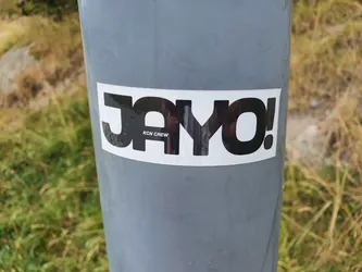 A black and white sticker with the word "Jayo!" in a bold, stylized font.  The text "KCN Crew" is smaller underneath. The sticker is affixed to a gray pole outdoors.
