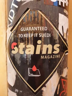 Black diamond-shaped sticker for Stains Magazine. It has a simple design with the magazine's name and a tagline. A small red blood drop and a stack of papers are below the text.