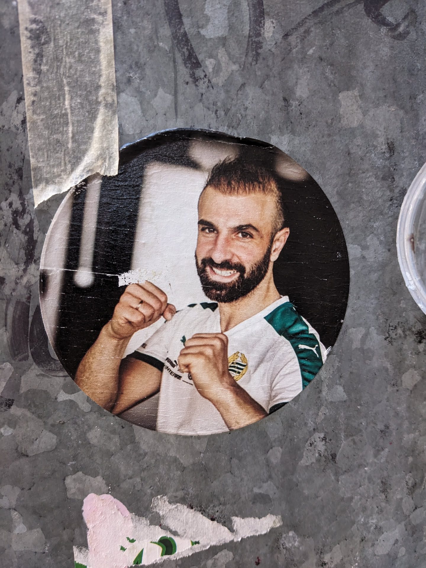 A round sticker featuring a close-up photo of a man with short brown hair and a beard. He's wearing a white and teal sports jersey with a logo on the chest and is shown in a fighting pose, fists clenched. The sticker is affixed to a textured grey surface.