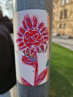 A sticker featuring a stylized flower with a red body and purple outlines. The flower has a face-like center. The sticker is affixed to a gray metal pole outdoors, with grass visible in the background.