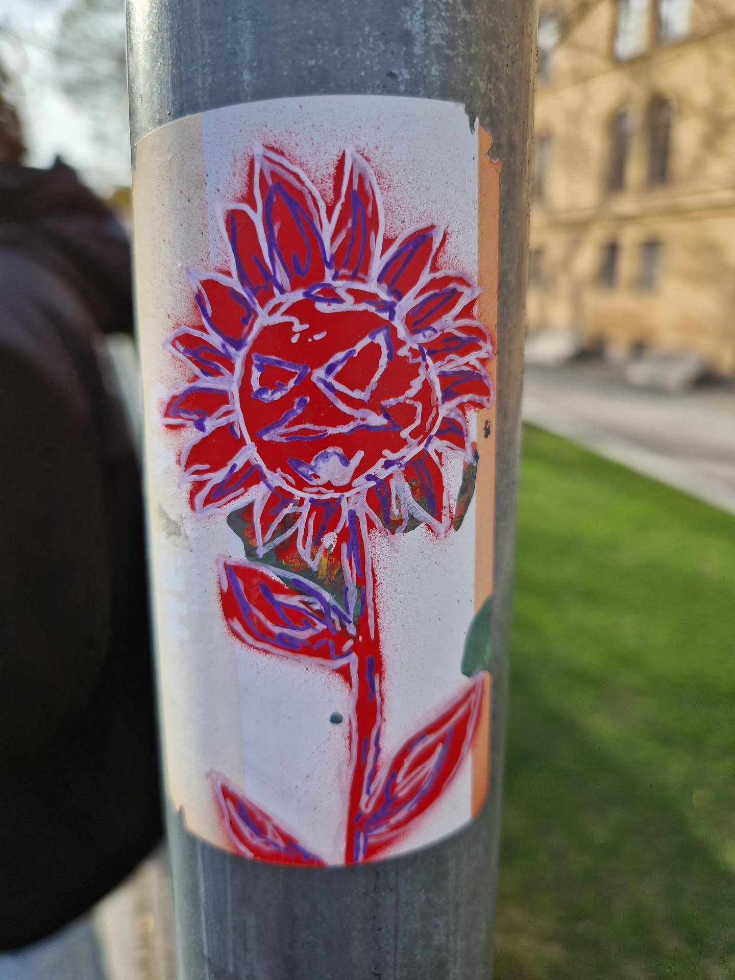 A sticker featuring a stylized flower with a red body and purple outlines. The flower has a face-like center. The sticker is affixed to a gray metal pole outdoors, with grass visible in the background.