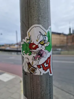 A sticker featuring a terrifying illustration of a clown with sharp teeth and green hair.  The style is reminiscent of cartoon horror. Red lettering is partially visible on the sticker. 