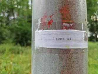 A small white sticker on a grey pole with the text "I sinom tid" written in typewriter style font. The pole is outdoors and surrounded by green vegetation.