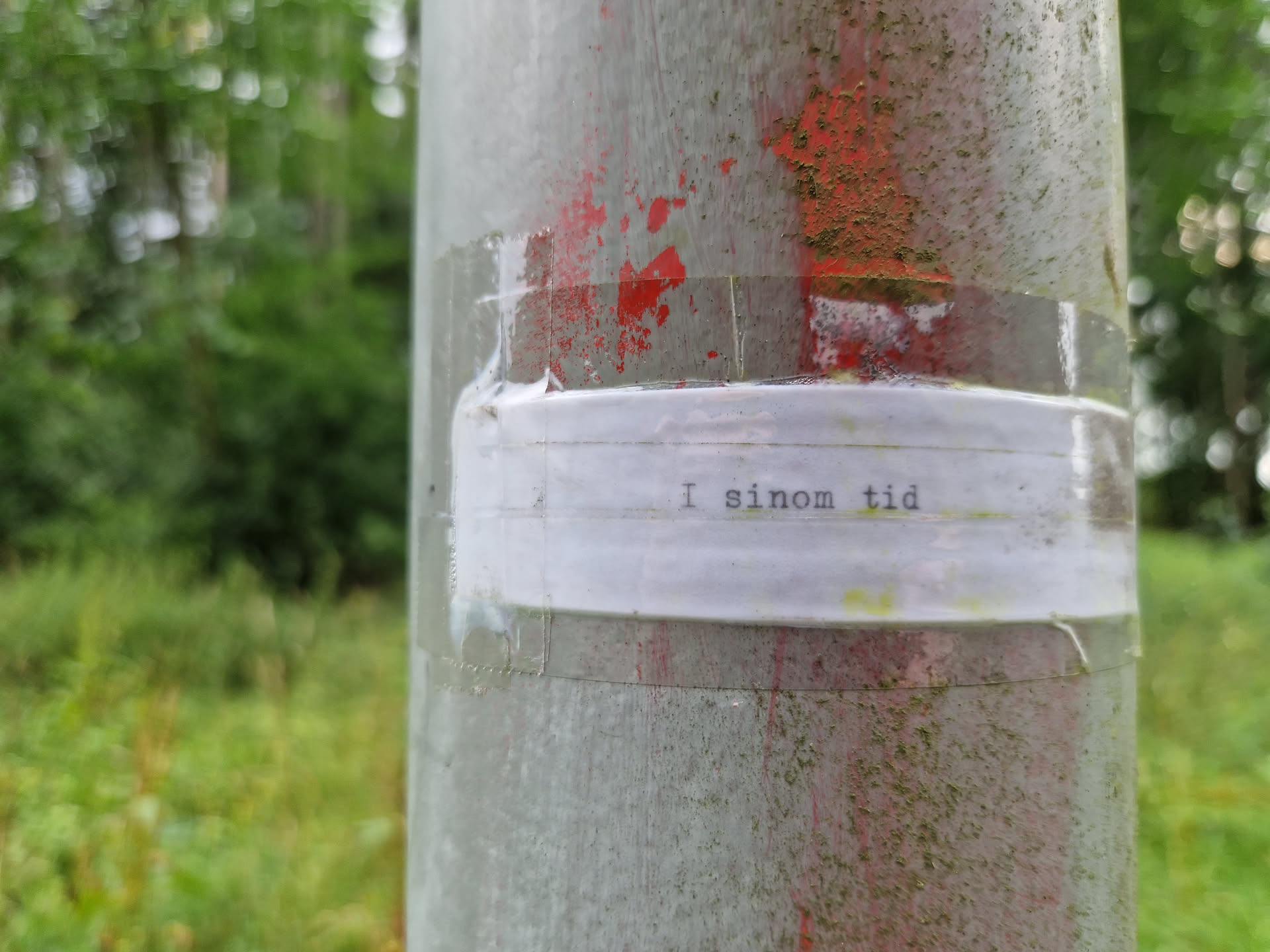 A small white sticker on a grey pole with the text "I sinom tid" written in typewriter style font. The pole is outdoors and surrounded by green vegetation.