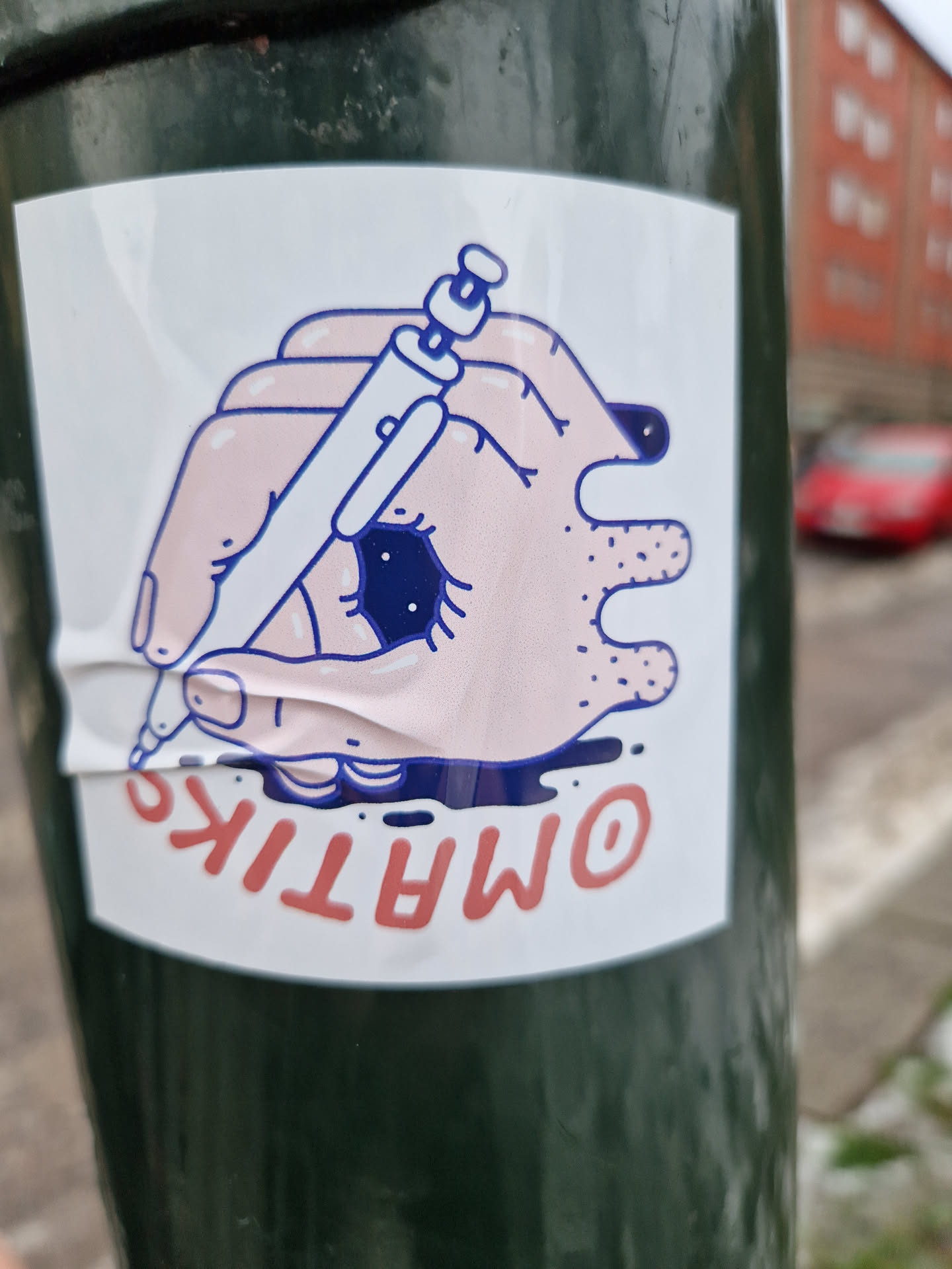 A sticker featuring a cartoon hand holding a pen and drawing near an eye. The text "OMATIK" is written underneath. 