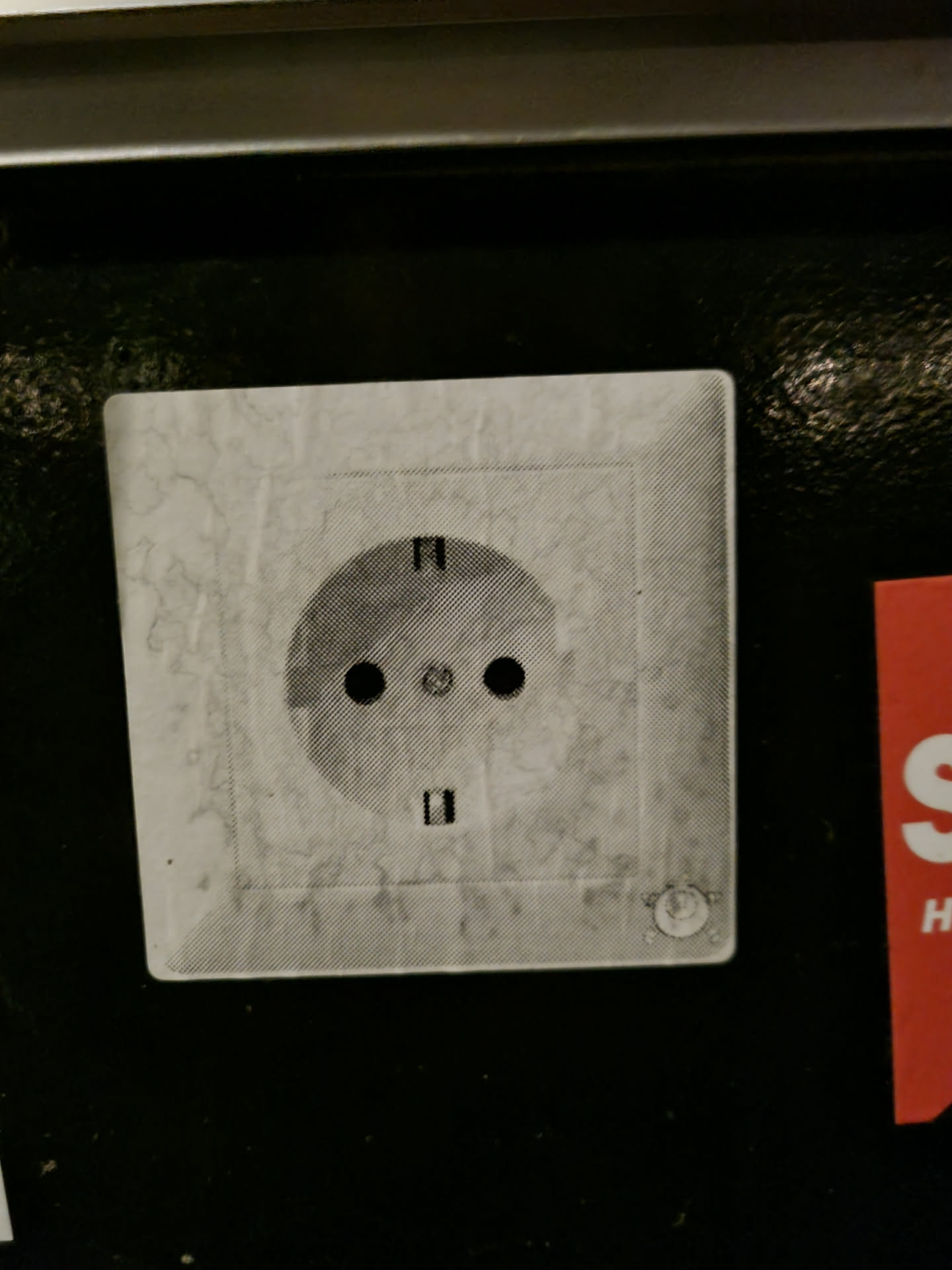 A slightly dirty and worn sticker depicting a European-style electrical power outlet. The sticker is affixed to a dark surface. 