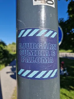 A sticker featuring the text 'DJURGÅRDEN CUMBIA & PALOMA' in a stylized font. The sticker has a dark blue background with light blue diagonal stripes at the top and bottom.