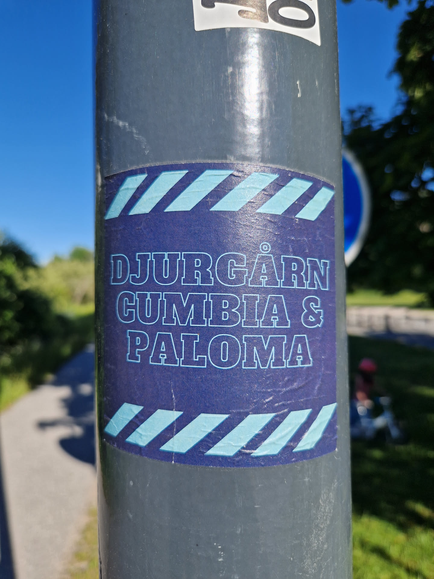 A sticker featuring the text 'DJURGÅRDEN CUMBIA & PALOMA' in a stylized font. The sticker has a dark blue background with light blue diagonal stripes at the top and bottom.