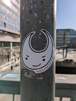 A white die-cut sticker featuring a cartoonish character with a crescent moon-like shape, a smiling face, and the text "SOUL COLLECTORS INC.". The sticker is adhered to a weathered metal pole, with an urban background featuring buildings and infrastructure.