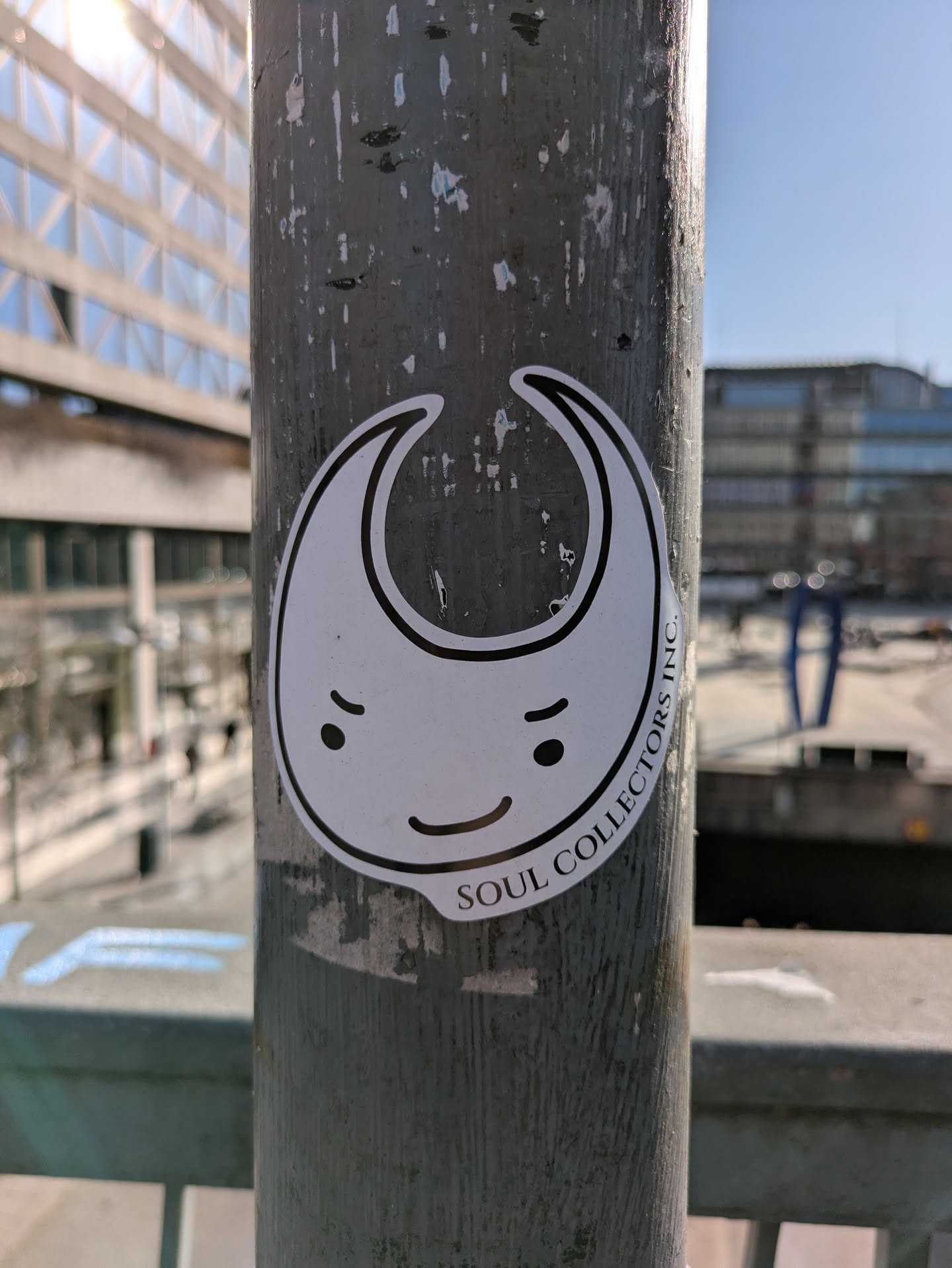 A white die-cut sticker featuring a cartoonish character with a crescent moon-like shape, a smiling face, and the text "SOUL COLLECTORS INC.". The sticker is adhered to a weathered metal pole, with an urban background featuring buildings and infrastructure.