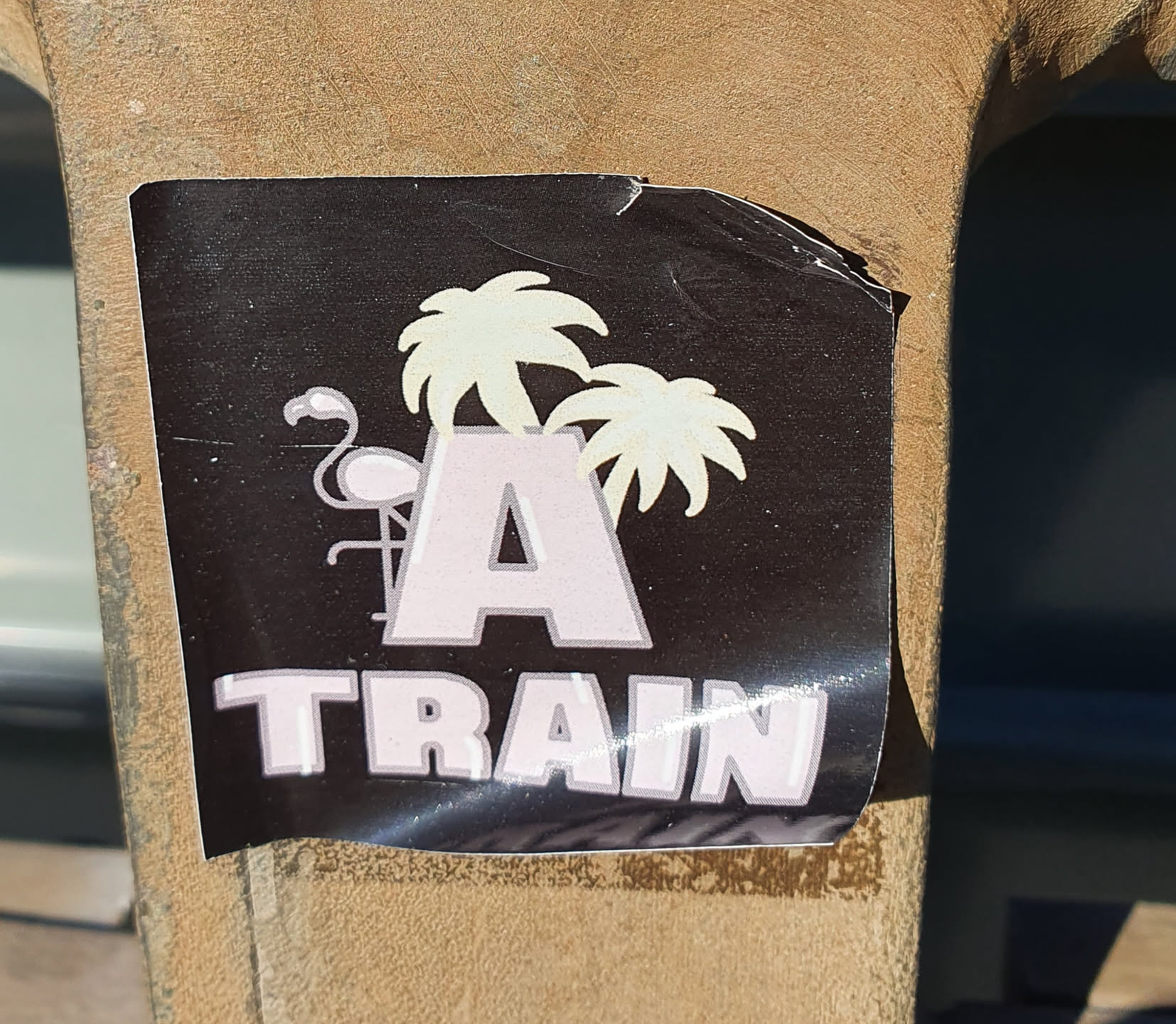A black sticker with a large letter A, palm trees, and a flamingo. The word "TRAIN" is written below the letter A. The sticker has a slightly worn appearance.