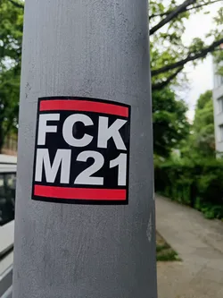 A square sticker with the text "FCk M21" in white on a black background. The sticker has a red stripe at the top and bottom. The sticker is affixed to a gray pole. 