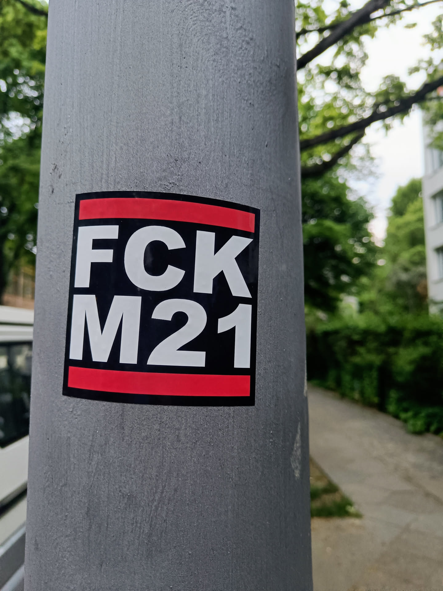 A square sticker with the text "FCk M21" in white on a black background. The sticker has a red stripe at the top and bottom. The sticker is affixed to a gray pole. 