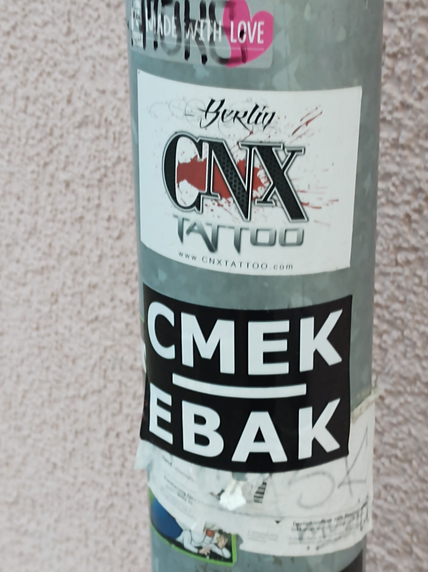 Black and white sticker with the text CMEK above a line and EBAB below.  It's stuck on a pole with other stickers, one of which is an advertisement for CNX Tattoo in Berkely. 