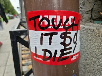 A red name tag sticker with black marker writing on it. The text says "TOUCH IT$D I DIE". It is stuck on a brown pole.