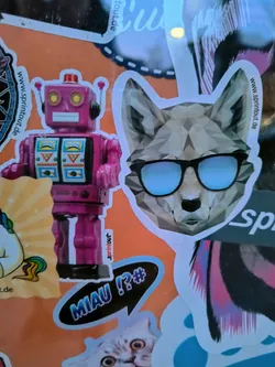 A sticker featuring a polygonal wolf wearing sunglasses. The style is geometric and minimalist.  There is also a pink robot sticker nearby and other various stickers. The background appears to be an orange surface.