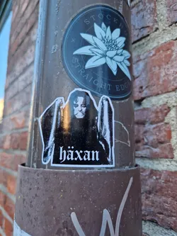 A black and white sticker featuring a figure with the word "Häxan" underneath. The sticker is affixed to a brown metal pole on a brick wall. There is another sticker above this one featuring a water lily with the words "Stockholm Straight Edge".