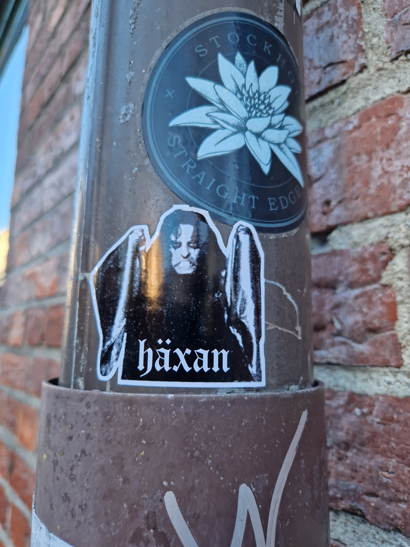 A black and white sticker featuring a figure with the word "Häxan" underneath. The sticker is affixed to a brown metal pole on a brick wall. There is another sticker above this one featuring a water lily with the words "Stockholm Straight Edge".