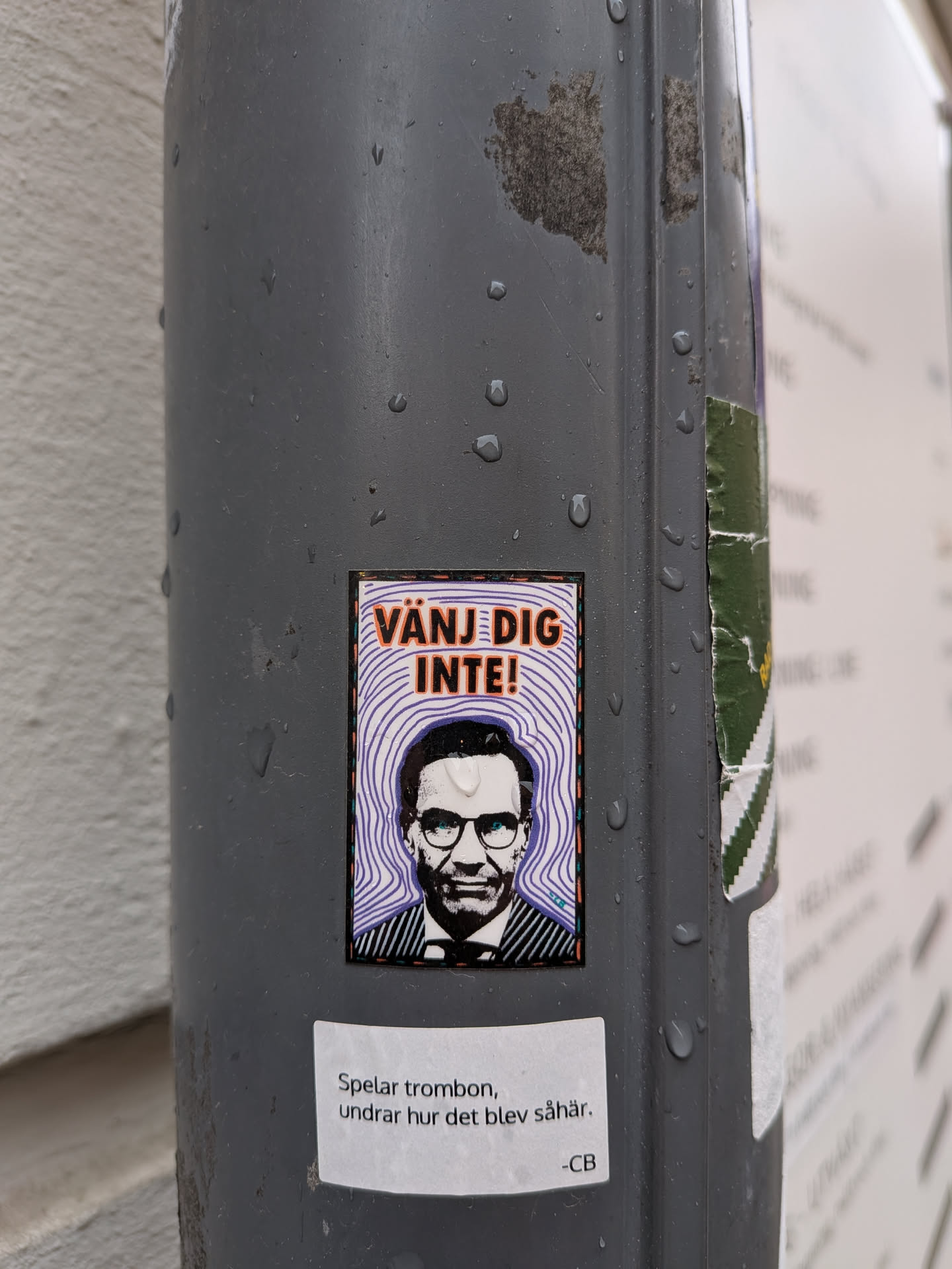 A rectangular sticker featuring a stylized, black and white portrait of a man, identifiable as Stefan Löfven, from the chest up. He wears glasses, a suit jacket, and a striped shirt with a tie, and has a serious expression. Behind his head, a background of concentric, wavy purple lines on a white base creates a dynamic effect. Above his head, the text 'VÄNJ DIG INTE!' is prominently displayed in bold orange capital letters with a black outline. The sticker is framed by a thin black border, an orange border, and a thicker black border adorned with small white dots. It is affixed to a wet, dark grey pole.