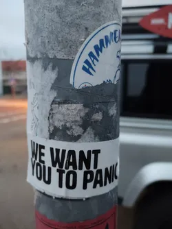A sticker on a pole with the text 'We Want You To Panic' in bold black letters on a white background. There is a partially visible sticker above it, with the word 'Hammer' written in blue.