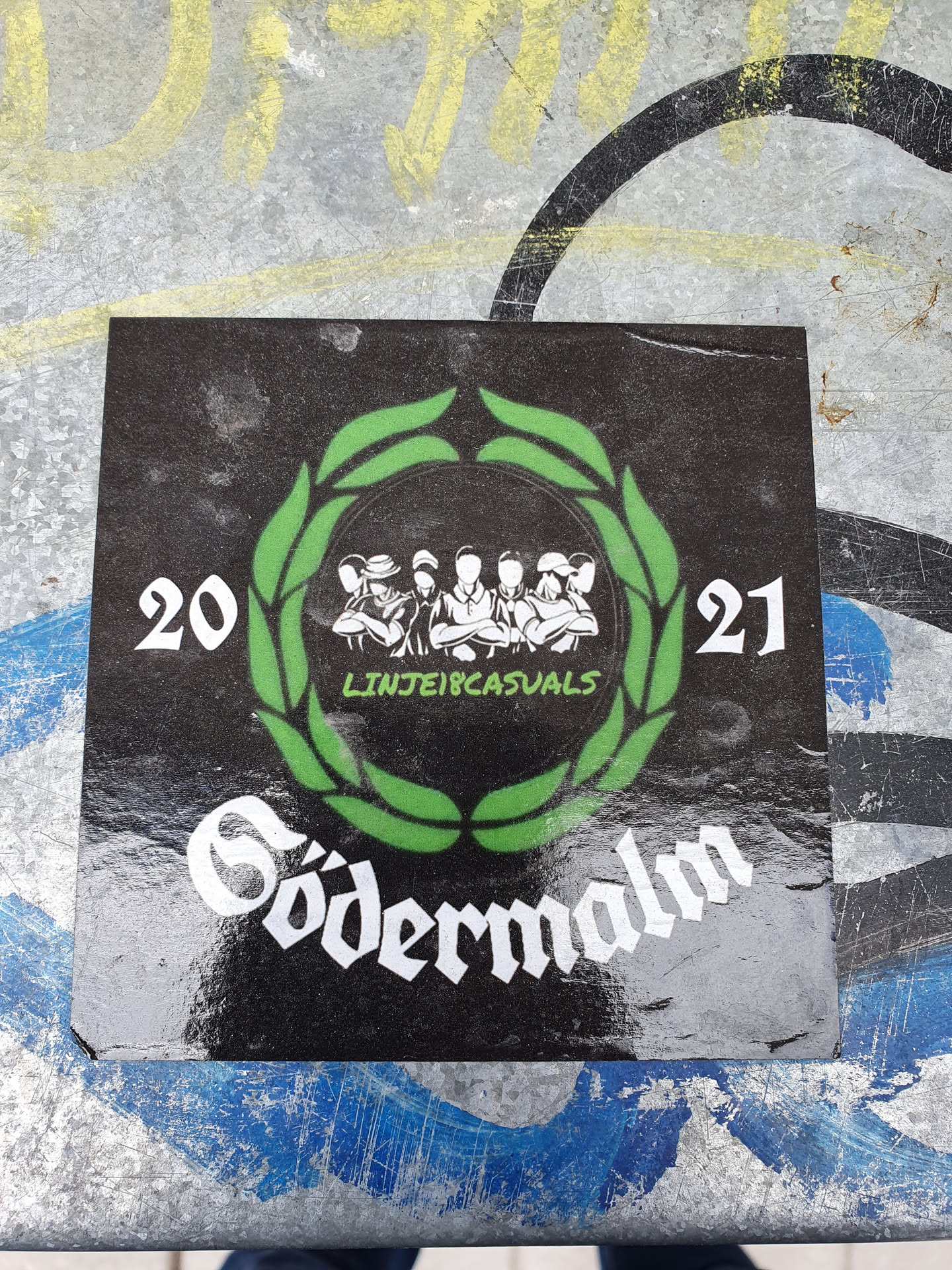 Black and white sticker with a green laurel wreath encircling a group of people. The text "LINJE18CASUALS" is visible inside the wreath. The year 20 and 21 are on the sides of the wreath and the word "Södermalm" is written in a stylized font at the bottom. 