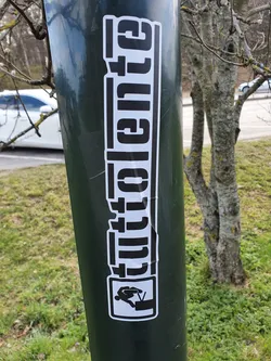 A black and white sticker featuring the word "Glitchente" in a stylized font, accompanied by a small graphic of a person jumping over a television. The sticker is affixed to a green pole outdoors.