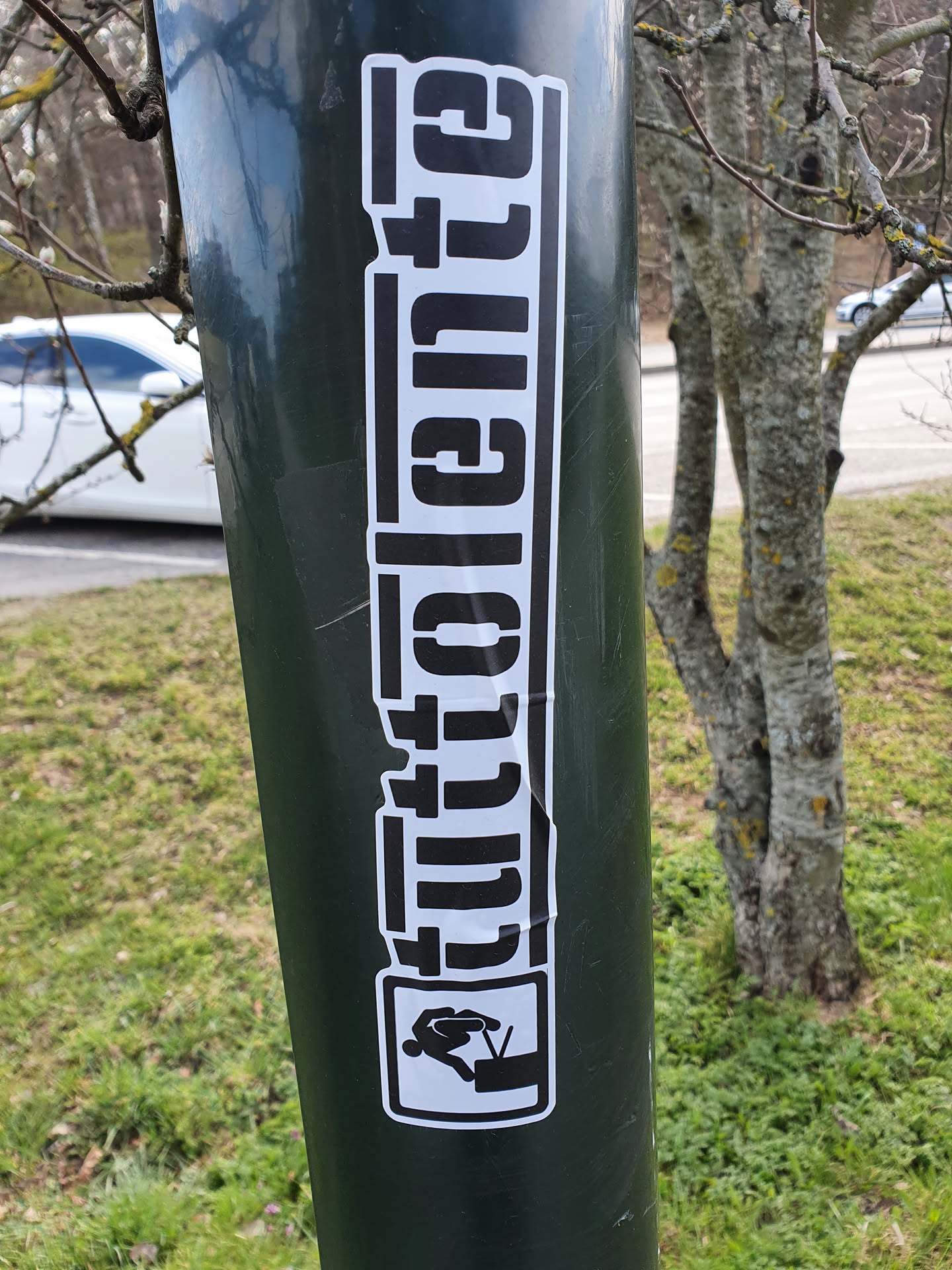 A black and white sticker featuring the word "Glitchente" in a stylized font, accompanied by a small graphic of a person jumping over a television. The sticker is affixed to a green pole outdoors.