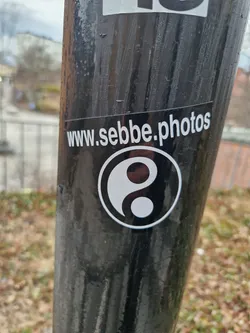 A black and white yin and yang symbol sticker with the website www.sebbe.photos printed below it. The sticker is affixed to a dark gray pole.