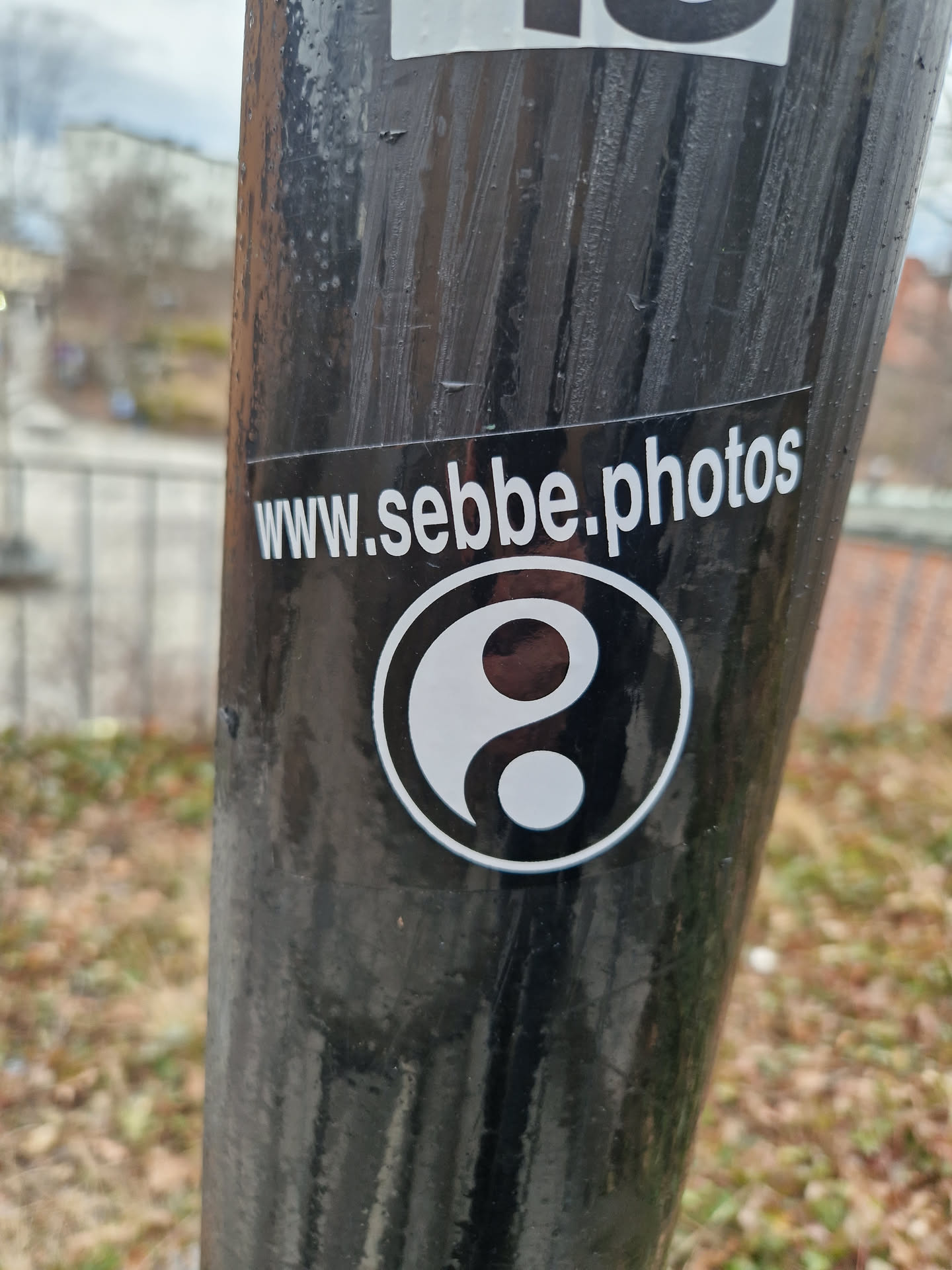 A black and white yin and yang symbol sticker with the website www.sebbe.photos printed below it. The sticker is affixed to a dark gray pole.
