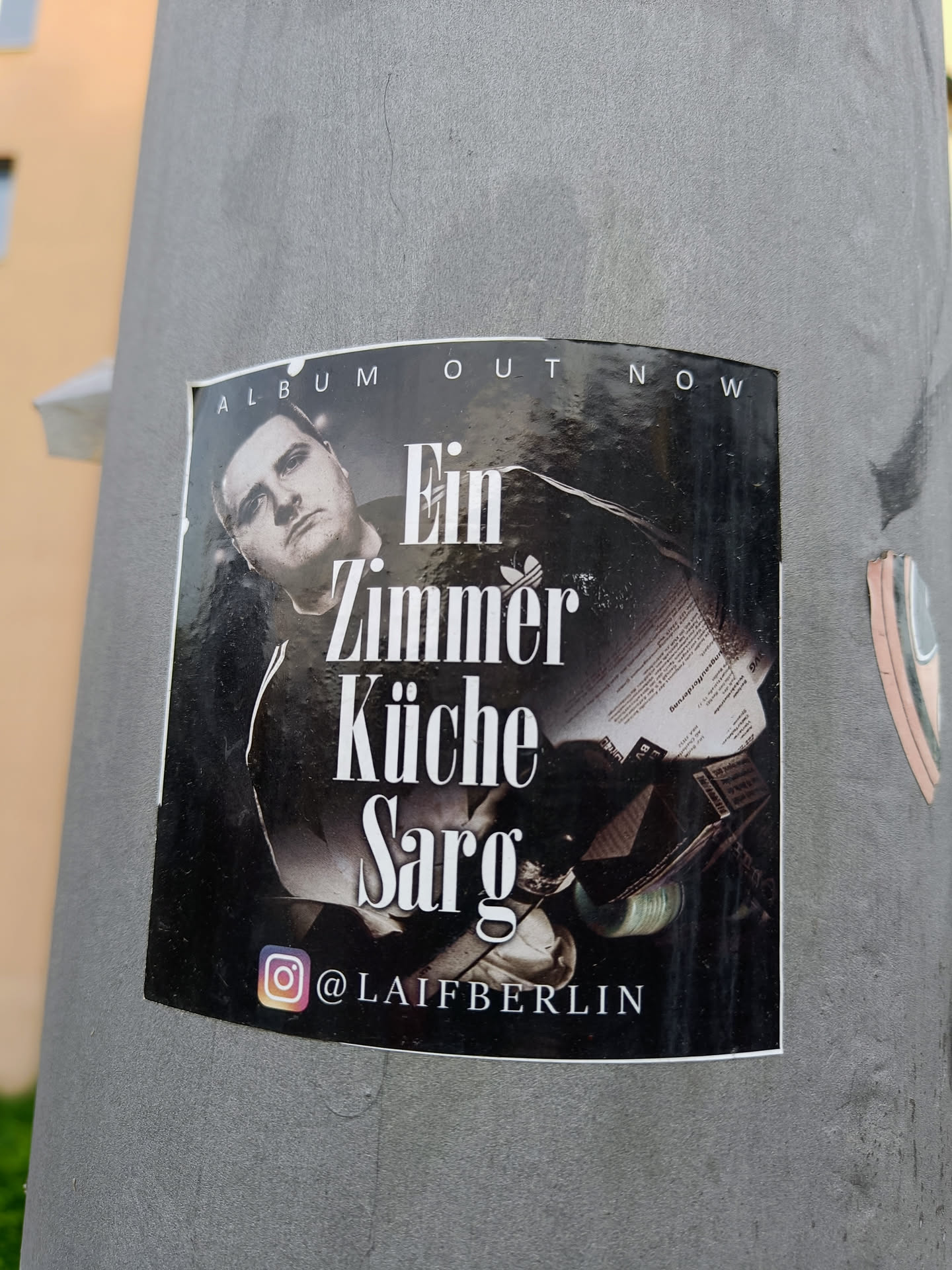 Black and white photo of a man on a sticker. The text on the sticker says "Album OUT NOW" at the top, and "Ein Zimmer Küche Sarg" in the center. There is an Instagram handle at the bottom: @LAIFBERLIN.